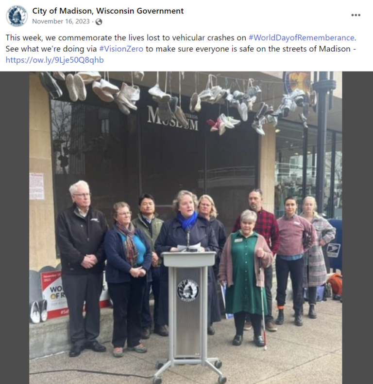 A group of people standing in front of a podium during a public event to raise awareness about road safety and the lives lost due to crashes. Above the podium, a rope is strung across the space and miltiple pair of shoes are tied to the string, hanging down in a row.