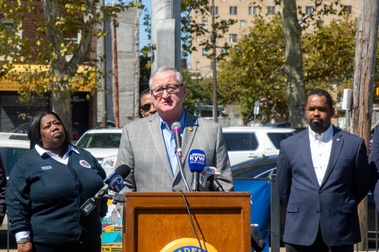 A mayor stands at a podium outdoors, speaking into microphones during a press conference. A parking lot is visible in the background.