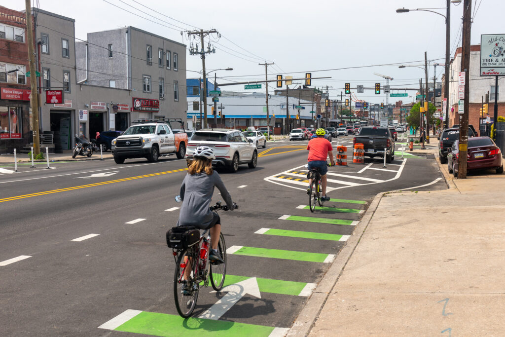 Cyclists riding on a green painted bike lane alongside a street with cars visible in the adjacent lanes