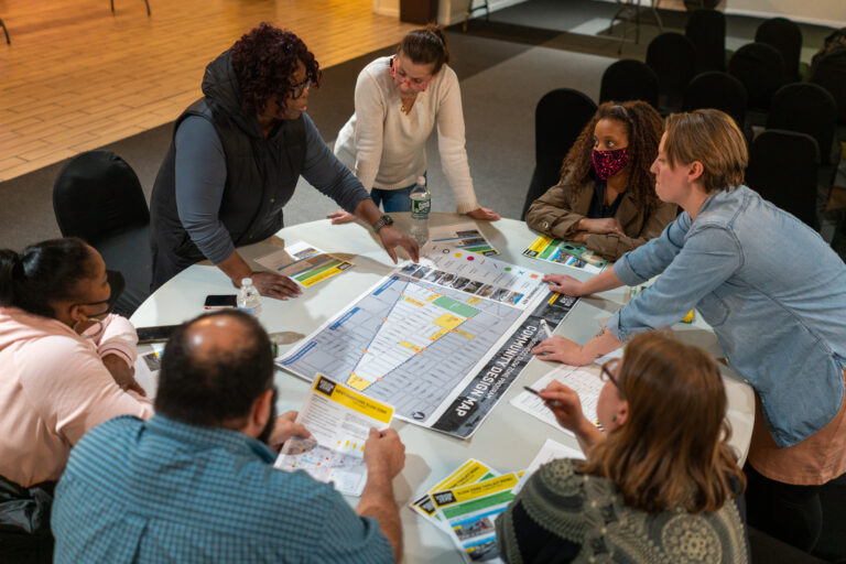 A group of people sitting around a table leaning in to examine a large map spread out on the table. The map shows an enlarged layout of a city block with street names. There are scattered flyers with information about slow zones on the table.