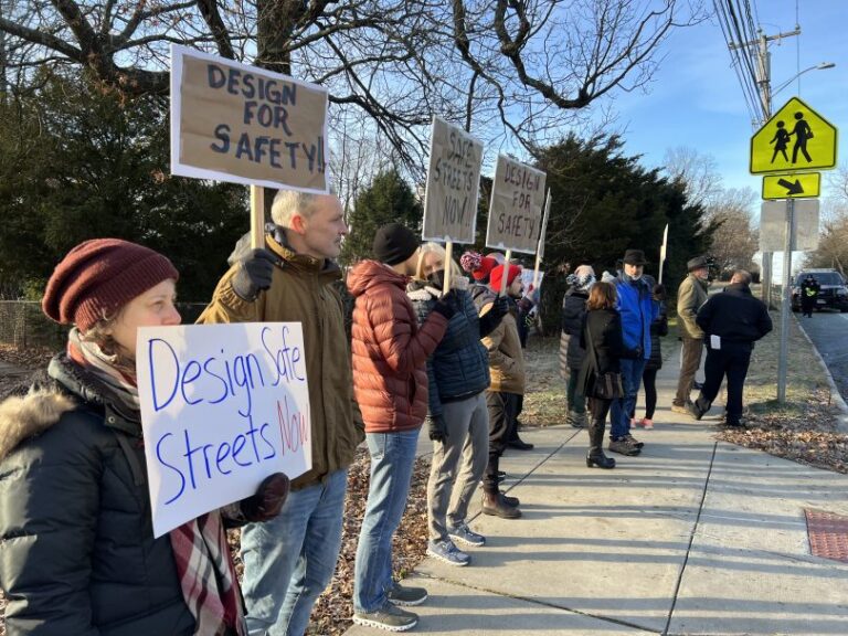 A group of people holding signs with slogans like " Design safe streets now", "Design for safety". They stand together in a sidewalk expressing their demand for safer streets.